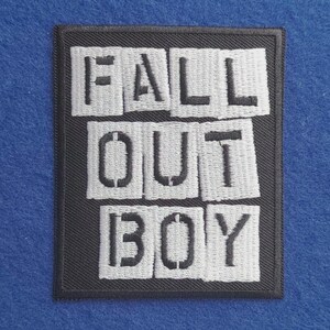 May include: A rectangular embroidered patch with a black border and a black background. The patch features the text FALL OUT BOY in white, stencil-style letters arranged in three rows, with each letter appearing on a separate white rectangular block.