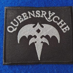 May include: A rectangular black embroidered patch featuring the silver-colored text "QUEENSRŸCHE" arched over a stylized silver winged emblem with a central diamond shape and a pointed base, all bordered by a thick black stitched edge.