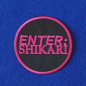 Può includere: Una toppa ricamata circolare con sfondo nero e bordo rosa brillante. Il testo ENTER: SHIKARI è ricamato al centro con un carattere audace e rosa brillante. La toppa presenta una trama in tessuto visibile e bordi cuciti ben definiti.