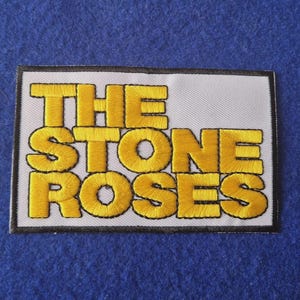 Op de afbeelding: Een rechthoekige geborduurde patch met de tekst THE STONE ROSES in dikke gele hoofdletters. De letters staan op een witte achtergrond en zijn omrand door een dunne zwarte lijn, wat zorgt voor een duidelijk en contrastrijk ontwerp.