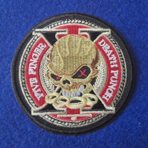 May include: A circular embroidered patch featuring a tan skull with red cross eyes centered over a black letter X. The design includes a red circular border with the white text FIVE FINGER DEATH PUNCH and a chain graphic at the bottom.