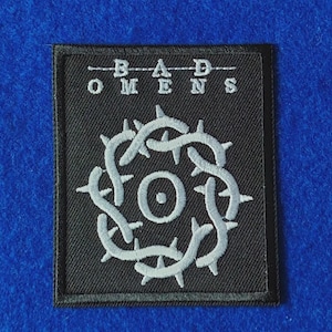 Puede incluir: Un parche rectangular negro bordado con el texto BAD OMENS en letras mayúsculas blancas en la parte superior. Debajo del texto hay un diseño circular blanco que se asemeja a una corona de espinas con un pequeño círculo sólido en el centro.