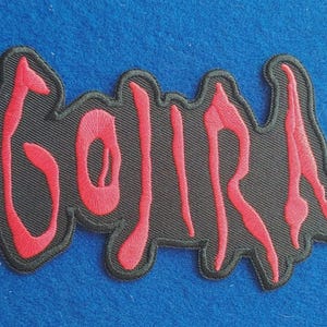 May include: An embroidered patch featuring the word GOJIRA in red thread on a black background. The lettering has a stylised, jagged font design with a thick black border outlining the entire shape of the text, creating a distinct contrast.