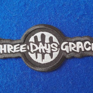 May include: A black and white embroidered patch featuring a circular emblem with a stylised graphic design in the centre. The text THREE DAYS GRACE is stitched in white lettering across the horizontal bar of the patch.