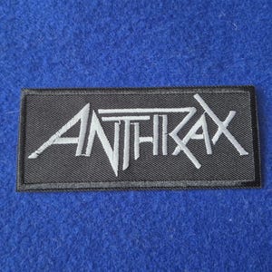 May include: A rectangular embroidered patch featuring a black background with the word ANTHRAX stitched in white thread. The lettering is stylised with sharp, angular lines and a distinct font design, finished with a black stitched border.