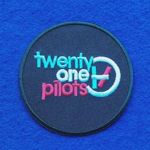 May include: A circular embroidered patch with a black background and a dark border. The text twenty one pilots is stitched in light blue and pink thread. A white circular logo with a pink diagonal line is positioned to the right of the text.