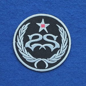 Op de afbeelding: Een ronde geborduurde patch met een zwarte achtergrond en een witte rand. Het ontwerp bevat een wit gestileerd dubbel S-monogram, een witte ster met een klein roze rechthoekje in het midden en witte lauriertakken.