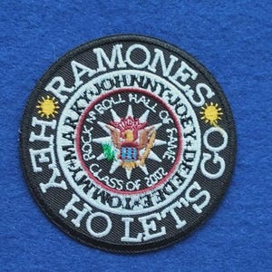 Può includere: Una toppa ricamata circolare con sfondo nero e bordo bianco. Il testo recita RAMONES, HEY HO LET'S GO, MARKY, JOHNNY, JOEY, DEE DEE, TOMMY, ROCK N ROLL HALL OF FAME e CLASS OF 2002. Presenta un emblema centrale con aquila e due soli gialli.
