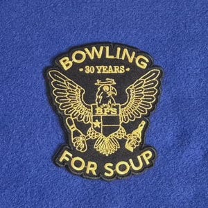 Puede incluir: Un parche bordado negro con un águila dorada de alas extendidas que sostiene un bolo de bolos y una botella. El texto dice BOWLING 30 YEARS FOR SOUP con BFS en un escudo. El parche está colocado sobre un fondo de tela azul sólido.