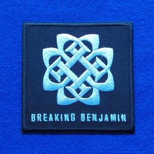 May include: A square black embroidered patch featuring a light blue Celtic-inspired knot design centred above the text BREAKING BENJAMIN in a matching light blue font. The patch has a stitched border and is set against a bright blue background.