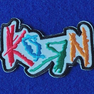 Op de afbeelding: Een geborduurde patch met de tekst KSYN in een gestileerd lettertype. De letter K is rood, de S is blauw, de Y is groen en de N is oranje. Het hele ontwerp is omlijnd met zwart garen op een lichtblauwe achtergrond.