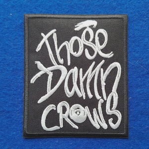 以下が含まれることがあります： 「Those Damn Crows」という白い刺繍文字がスタイリッシュな筆記体で施された、長方形の黒い刺繍ワッペン。ワッペンにはきれいな縁取りが施されており、布製品に装飾として縫い付けたり貼り付けたりできるようにデザインされています。