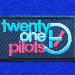 May include: A rectangular embroidered patch with a black background. The text twenty one pilots is stitched in light blue and pink thread. A circular logo featuring a light blue ring and a pink diagonal slash is positioned on the right side.