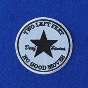 Op de afbeelding: Een ronde geborduurde patch met een witte achtergrond en een zwarte rand. Een grote zwarte ster staat in het midden. De tekst "TWO LEFT FEET" staat in een boog bovenaan, "Deaf" en "Havana" flankeren de ster en "NO GOOD MOVES" staat onderaan.