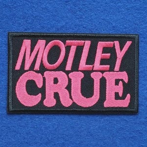 May include: A rectangular embroidered patch with a black background and a black stitched border. The text MOTLEY CRUE is embroidered in a bold, bright pink font across two lines, with MOTLEY on the top line and CRUE on the bottom line.