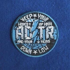 May include: A circular embroidered patch featuring a light blue lightning bolt in the centre. The text reads: KEEP YOUR HOPES UP HIGH, AND YOUR HEAD DOWN LOW, with the letters ADTR prominently displayed in white and black stitching.