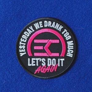 May include: A circular embroidered patch with a black background. It features a pink stylised logo in the centre and white text that reads YESTERDAY WE DRANK TOO MUCH and LET'S DO IT AGAIN in pink script at the bottom of the circular design.