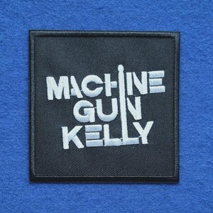 Op de afbeelding: Een vierkante zwarte geborduurde patch met de witte tekst "MACHINE GUN KELLY" in een vet, blokkerig lettertype. De letter "I" in "MACHINE" is gestileerd als een verticaal luciferstokje. De patch heeft een zwarte gestikte rand.