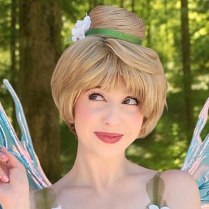 May include: A blonde wig styled in a high bun with bangs, accented by a green headband and white flowers. The costume features a green bodice adorned with layered leaf-like fabric and white floral appliqués, paired with large iridescent blue wings.