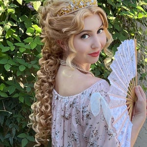 May include: A gold-toned metal tiara with clear crystals, a multi-strand pearl necklace, and a white lace folding fan with wooden ribs. The ensemble is paired with a light pink dress featuring a dark floral print and white ribbon shoulder ties.