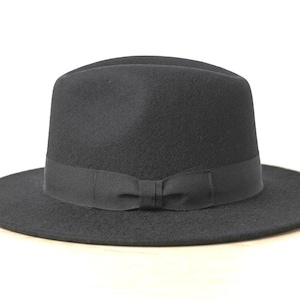 May include: A solid black fedora hat with a structured crown and a wide, flat brim. The hat features a matching black grosgrain ribbon band encircling the base of the crown, which is finished with a small, centred bow detail on the front.