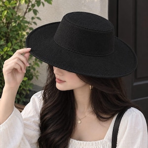 May include: A black hat with a flat crown and a wide, structured brim. The hat features a textured, felt-like surface material and a clean, circular silhouette. The design includes a distinct seam where the crown meets the brim, providing a solid, uniform appearance in a deep, matte black colour.
