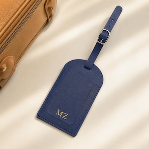 Pode incluir: Uma etiqueta de bagagem azul-marinho com acabamento texturizado e uma alça correspondente com fivela de metal prateado. A etiqueta tem formato retangular com cantos superiores arredondados e as letras douradas MZ gravadas na parte inferior.
