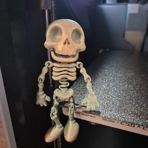 May include: A 3D printed articulated skeleton figurine with a pale, off-white finish and visible layer lines. The figure features a large rounded skull, a rib cage, and jointed limbs with hands and feet, positioned on a textured print bed.