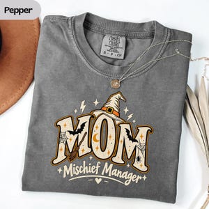 Peut inclure: Un t-shirt gris anthracite à manches courtes avec une impression graphique indiquant MOM Mischief Manager. Le motif comprend un chapeau de sorcière, un balai, des chauves-souris, des toiles d'araignée et des étoiles en orange, noir et crème.