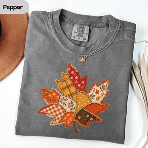 Könnte beinhalten: Ein anthrazitfarbenes Kurzarm-T-Shirt mit einer großen, zentralen Grafik eines Ahornblatts, das aus verschiedenen Patchwork-Mustern in Orange-, Rot-, Gelb- und Brauntönen besteht. Das T-Shirt hat einen Rundhalsausschnitt und ein Comfort Colors-Etikett.
