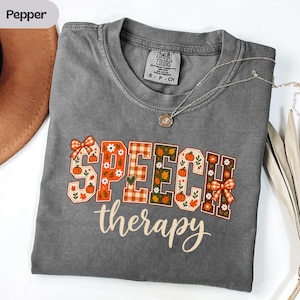 Könnte beinhalten: Ein anthrazitfarbenes T-Shirt mit dem Schriftzug "SPEECH therapy" in einer dekorativen Schriftart. Die Buchstaben sind mit herbstlichen Mustern gefüllt, darunter orangefarbene Kürbisse, weiße Blumen, grüne Blätter und orangefarbene Karomuster.