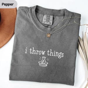以下が含まれることがあります： チャコールグレーの半袖クルーネックTシャツ。シャツには「i throw things」という白い文字と、ろくろと花瓶のシンプルな線画がプリントされています。細いゴールドトーンのチェーンネックレスが上に置かれています。