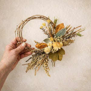 May include: A circular wreath constructed from thin, brown, intertwined twigs. One side of the wreath is adorned with a cluster of dried botanical elements, including yellow flowers, green leaves, brown foliage, and slender stalks in shades of tan.
