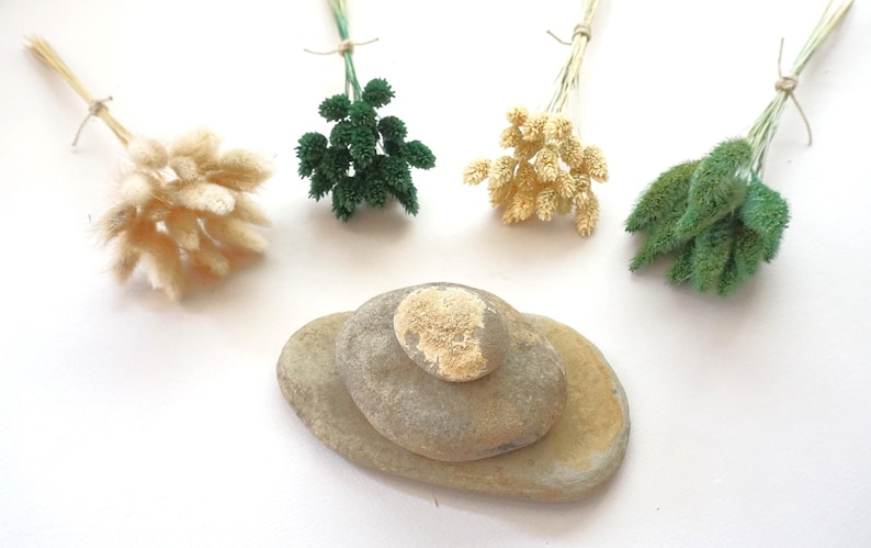 May include: Four small bundles of dried grass in cream, dark green, light yellow, and sage green colors, each tied with twine. Below the bundles are three smooth, flat, gray and tan river stones stacked in a pyramid formation on a white surface.