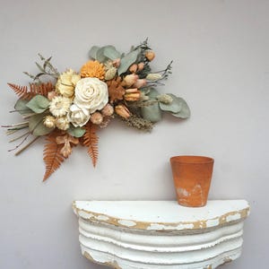 May include: A floral arrangement featuring cream, orange, and muted green dried flowers, including roses, ferns, and eucalyptus leaves. Below the arrangement sits a terracotta pot on a white, distressed, semi-circular wooden wall shelf.