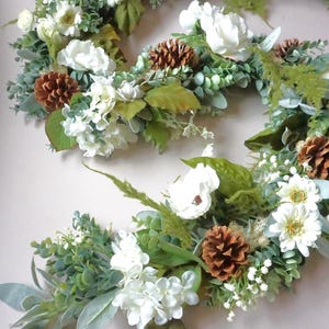 May include: A white and green floral garland with pine cones. The garland is made of artificial flowers and greenery.
