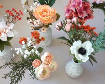 May include: Four small white textured ceramic vases each containing a unique arrangement of artificial flowers including peach roses, white anemones with black centers, orange berries, green foliage, and delicate white baby's breath blossoms.