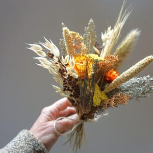 May include: A small bouquet of dried flowers and grains, held in a hand. The arrangement features a mix of beige, brown, yellow, and orange elements, tied together with twine. The background is a soft gray.