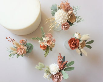 May include: Five small floral arrangements featuring dried and faux flowers in shades of rust, cream, and sage green, including roses, dahlias, and grasses, positioned around a large, smooth, circular white object on a light surface.