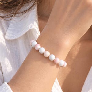 Può includere: Un braccialetto di perline con un motivo ripetuto di sfere lisce bianche e rosa tenue. Le perline sono di dimensioni uniformi e disposte in un unico filo, creando un design semplice e pulito che circonda il polso in modo regolare.