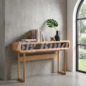 May include: A light-toned wooden console table with a rounded rectangular open shelf design. On top sits a stack of books, a gold geometric wire sculpture, a small wooden cross-shaped object, and a black vase containing a green monstera leaf.