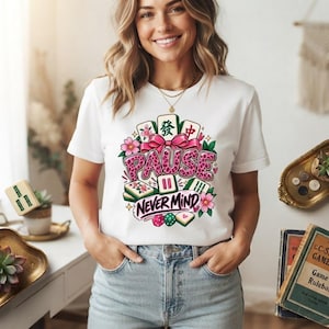 May include: A white short-sleeve t-shirt featuring a colorful graphic of mahjong tiles, pink cherry blossoms, and a pink bow. The text reads PAUSE in leopard print and NEVER MIND in purple script, surrounded by small dice and heart accents.
