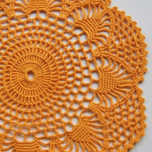 May include: A crocheted orange doily with a circular design. The doily has a delicate, intricate pattern with a central circle and radiating spokes.