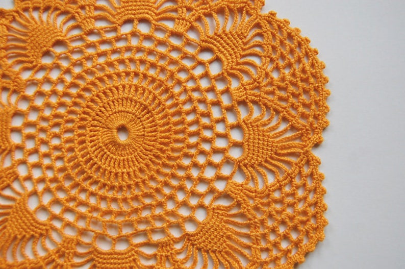 May include: A crocheted orange doily with a circular design. The doily has a delicate, intricate pattern with a central circle and radiating spokes.