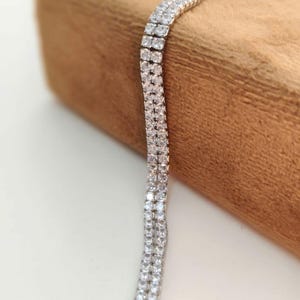 May include: A double-row tennis bracelet featuring two parallel lines of clear, round-cut stones set in a silver-toned metal chain. The bracelet displays a consistent, sparkling design with each stone securely held in a metallic prong setting.