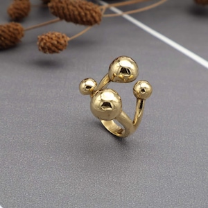 May include: A gold-toned ring featuring an open band design with four polished spherical accents of varying sizes. The metal surface is smooth and reflective, showcasing a rounded, multi-ball structure that creates a distinct, dimensional appearance.