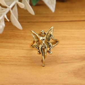 May include: A gold-toned ring featuring a central figure of a winged fairy with outstretched arms and legs positioned together. The fairy design is integrated into the band, with the wings extending outward to form the sides of the ring structure.