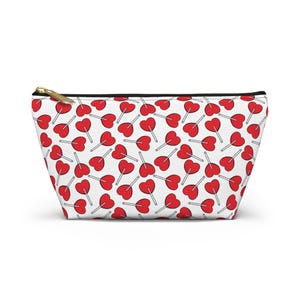 May include: A white zippered pouch featuring a repeating pattern of red heart-shaped lollipops with thin white sticks. The bag has a black zipper closure at the top and a gold-toned metal zipper pull, providing a functional storage solution.