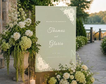 Puede incluir: Un letrero de bienvenida de boda rectangular de color verde salvia con ilustraciones florales blancas en las esquinas y el texto Welcome to the wedding of Thomas and Gloria 23.09.2024. El letrero se exhibe con arreglos florales blancos y velas.