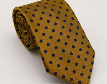 May include: A rolled necktie in a mustard yellow color featuring a repeating pattern of small, dark navy blue polka dots. The fabric has a visible diagonal weave texture, and the tie is folded into a coil at the top with a pointed end.